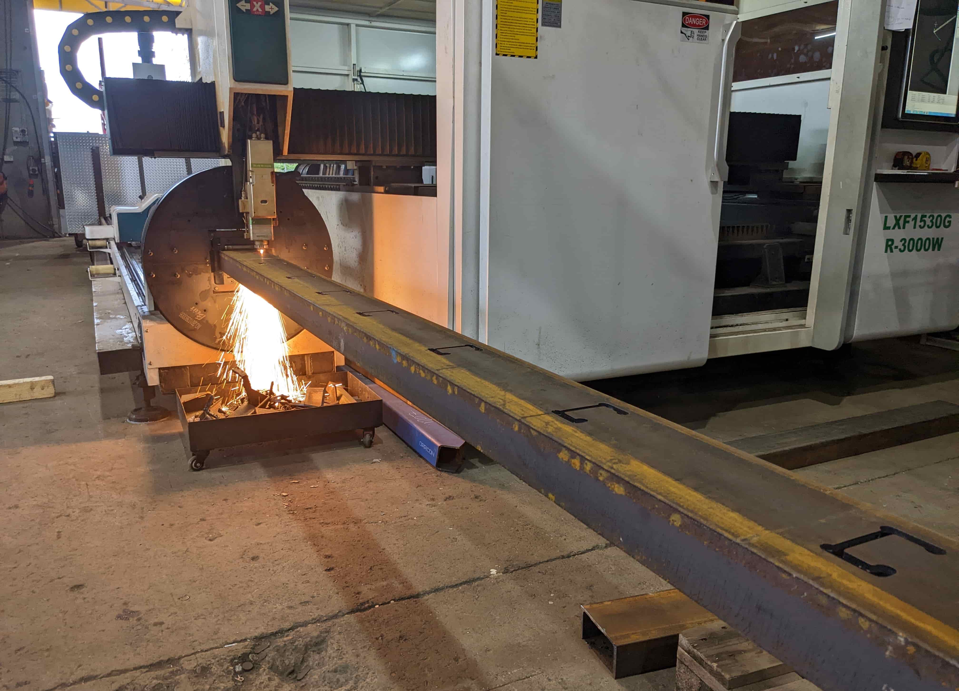Four-axis fibre laser cutting a long steel beam with sparks falling beneath the cutting bed