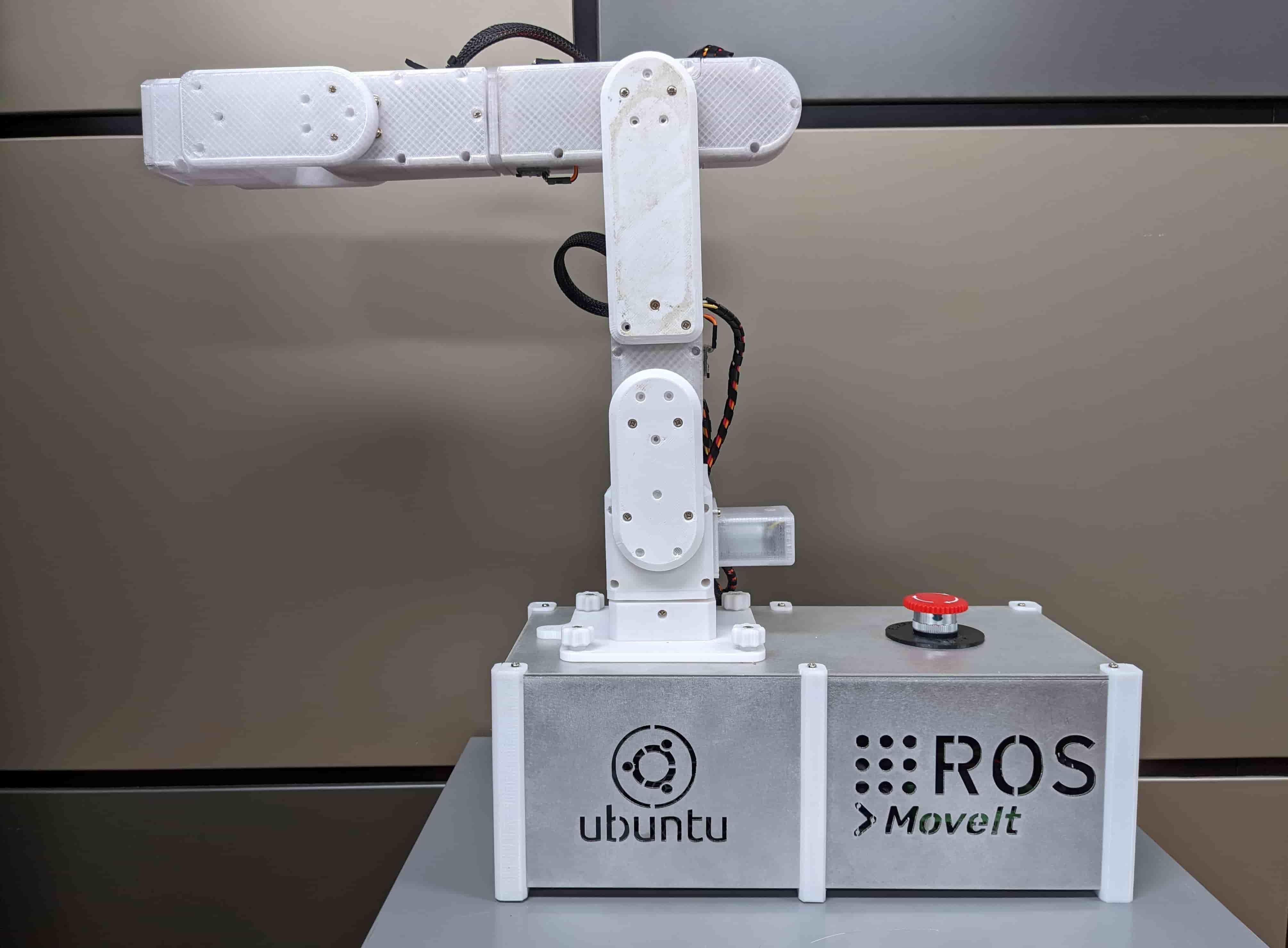 The finished six-axis arm — white 3D-printed links on a machined aluminium base carrying Ubuntu and ROS MoveIt marks, with a red emergency stop